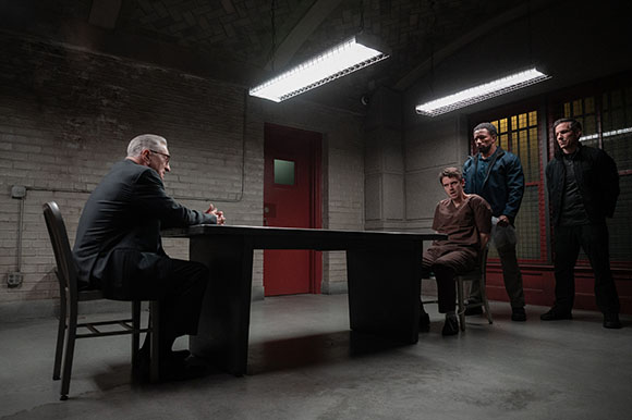 A man in a suit sits at a table facing three men in a dark, industrial room.