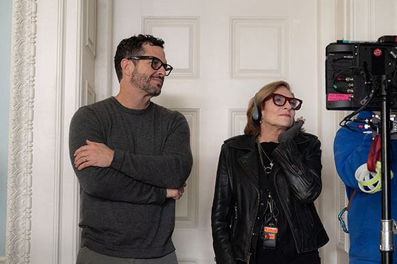 A man in a grey sweater and a woman in a black leather jacket stand near a professional film camera.