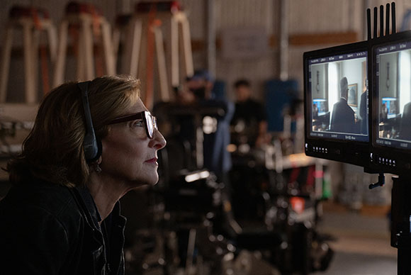A woman with glasses and headphones watches a film production monitor in a dimly lit studio.