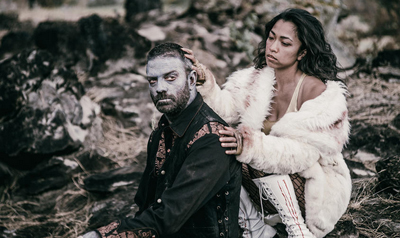 A woman in a white fur coat places her hand on a man with pale face paint sitting in a rocky landscape.