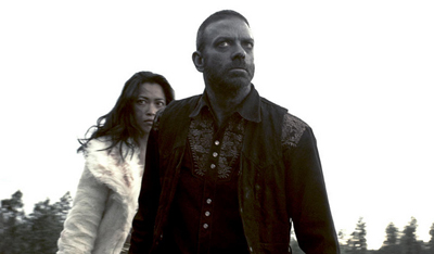 A man with a beard and a woman in a white coat look forward with serious expressions against a bright sky.