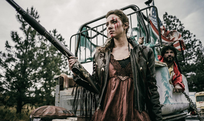A young woman with facial marks holds a long gun while standing in front of a decorated vehicle.