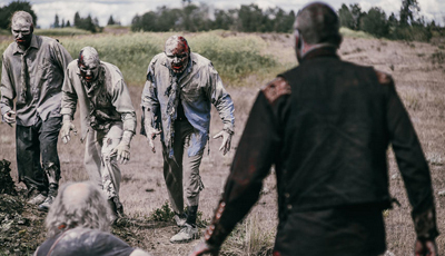 Four people in zombie makeup and tattered clothing walk through a dry field toward a person in a dark shirt.