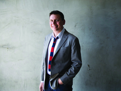 A smiling man wearing a grey suit jacket, white shirt, and red and blue striped tie stands against a grey wall.