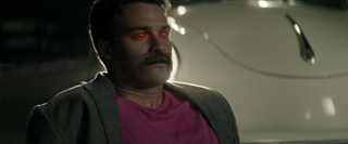A man with a mustache and glowing red eyes stands in front of a white car at night.
