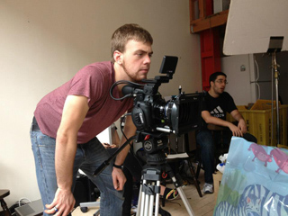 A man in a maroon shirt leans over a professional camera mounted on a tripod in a studio setting.