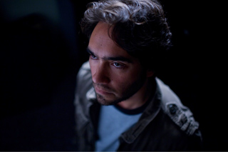 A man with dark hair and a beard looks pensively into the distance in dim lighting.