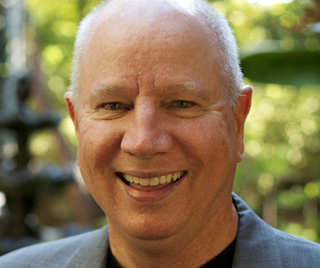 A smiling middle-aged man with short grey hair wearing a grey blazer against a blurred green background.