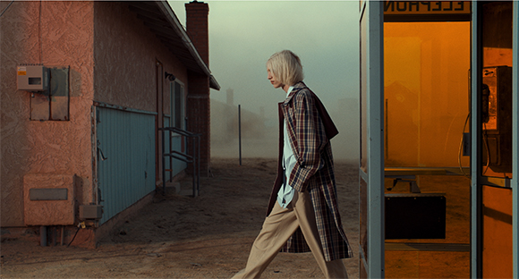 A person in a plaid coat walks past a pink building and an orange-lit telephone booth in thick fog.