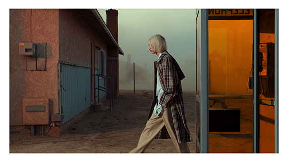 A person in a plaid coat walks through fog past a pink building and an orange telephone booth.