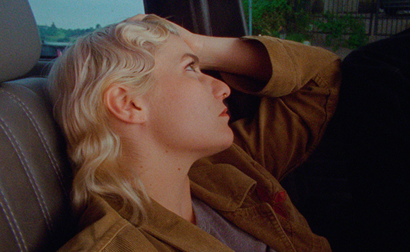 A blonde woman in a tan jacket leans her head against her hand while looking up inside a vehicle.