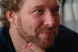Close-up of a man with light hair and a reddish-brown beard looking to the side with a slight smile.