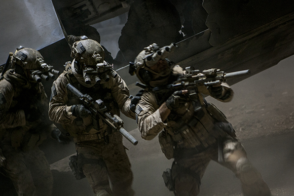 Three soldiers in camouflage and tactical helmets move forward through dust with rifles raised.