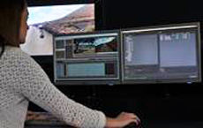 A person in a white patterned shirt sits before three computer monitors displaying various data and landscapes.