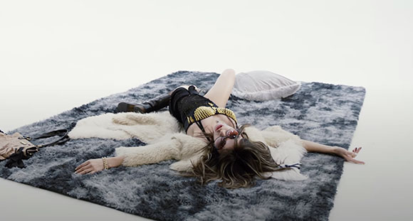 A person in a black top and white faux fur coat lies on a grey shag rug with sunglasses on.