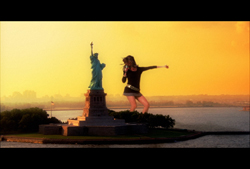 A person stands with arms outstretched next to the Statue of Liberty during a golden sunset.