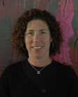 A woman with curly brown hair and a black shirt smiling in front of an abstract pink and grey painting.