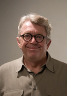 A smiling man with short light hair and round glasses wearing a tan button-down shirt.