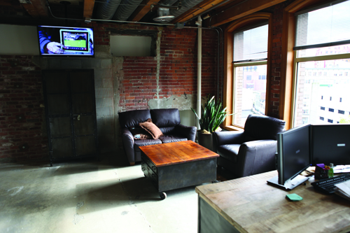 Industrial loft with brick walls, a dark leather sofa, a wooden coffee table, and large windows.