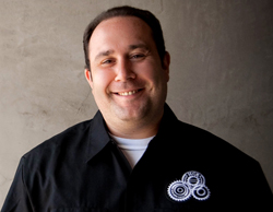 A smiling man wearing a black button-down shirt with a white gear logo on the chest.
