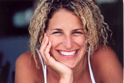 A smiling woman with curly blonde hair rests her chin on her hand.