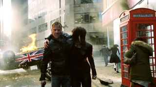 A man and woman walk through a smoky city street past a burning car and a red telephone booth.