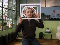 A man holds a white frame in front of his face, showing multiple reflections of himself inside the frame.
