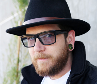 A man with a beard, black fedora, black-rimmed glasses, and a large ear gauge looking toward the camera.