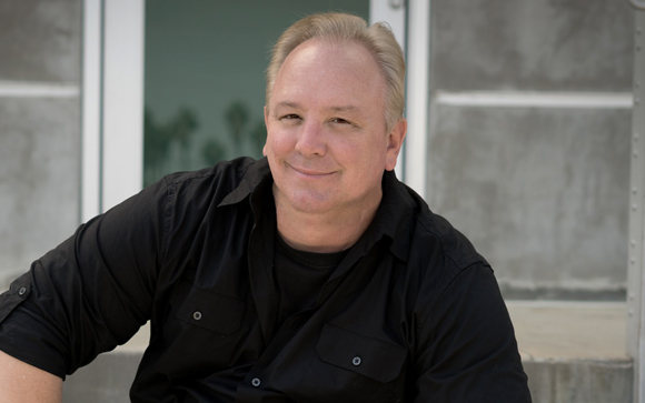 A man with light hair wearing a black button-down shirt, smiling at the camera.