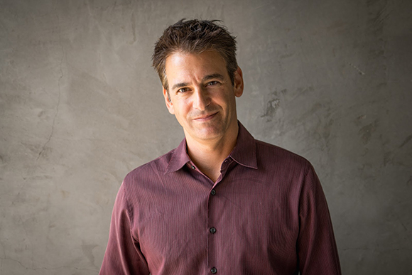 A man with short dark hair wearing a maroon button-down shirt, smiling against a textured gray background.