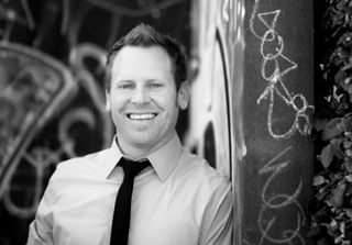 A smiling man wearing a collared shirt and tie stands in front of a graffiti-covered wall.