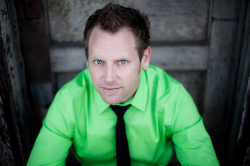 A man wearing a lime green button-down shirt and black tie looks upward against a dark wooden background.