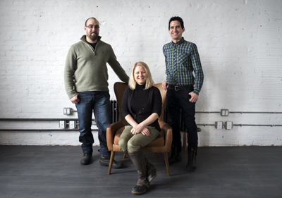 A man in a green sweater, a woman sitting in a brown chair, and a man in a plaid shirt stand against a white wall.