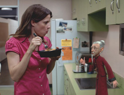 A woman in a pink shirt eats from a black bowl while looking at a small puppet on a kitchen counter.