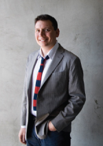 A smiling man wearing a grey blazer, white shirt, and red and blue striped tie standing against a grey wall.