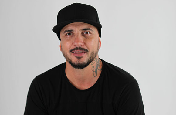 A man with a beard and neck tattoo wearing a black baseball cap and black t-shirt against a white background.