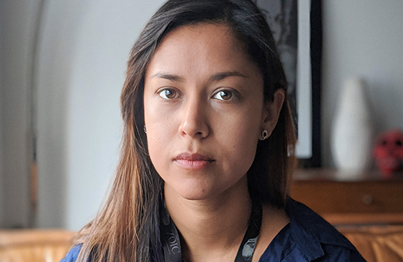 A woman with long brown hair looks forward with a neutral expression.