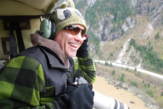 A man in a green plaid jacket and headset smiles while looking out of a helicopter over a river valley.
