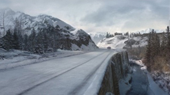 A snow-covered road stretches through a valley between rugged, snow-capped mountains under a cloudy sky.