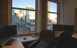 Office workspace with two desks, computer monitors, and chairs facing windows with a city view.