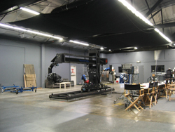 A large black camera crane on a track inside a spacious, dimly lit film studio.