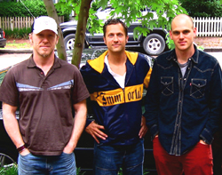 Three men standing side by side outdoors in front of a dark vehicle and green foliage.