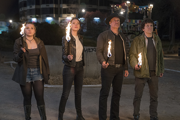 Four people stand in a line at night, each holding a flaming torch and looking upward.