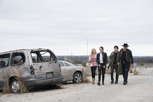 Four people walk past a damaged silver minivan and a white car in a barren, dusty field.