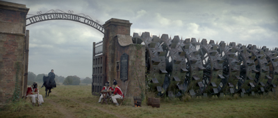 A massive spiked metal wheel structure sits beside a brick entrance gate under a cloudy sky.