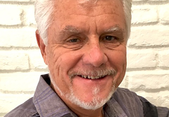 Close-up portrait of a smiling man with white hair and a white goatee against a white brick wall.