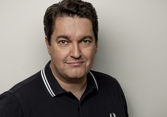 A man with dark wavy hair and a black polo shirt looking forward against a plain light background.
