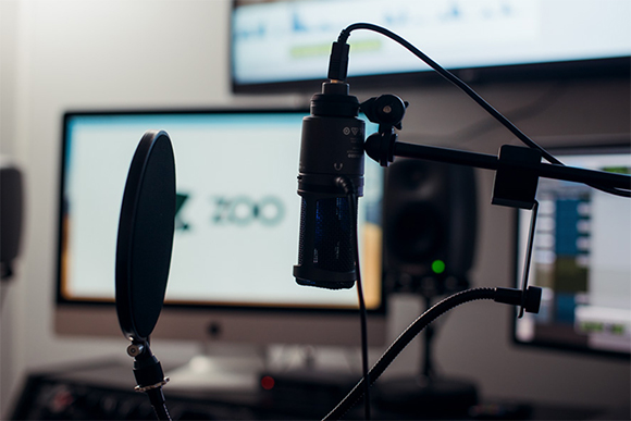A black condenser microphone with a pop filter in a studio with computer monitors in the background.