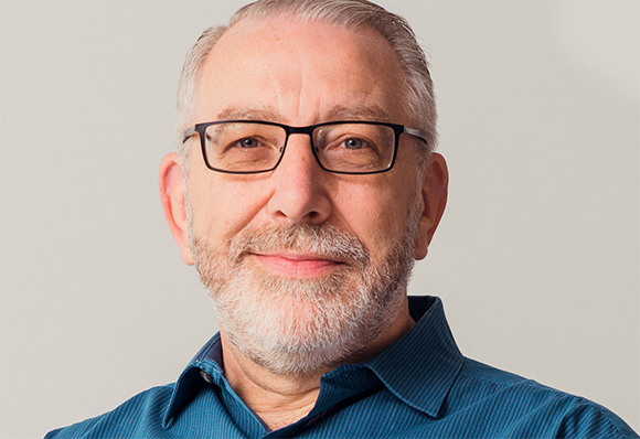 A smiling man with grey hair, glasses, and a white beard wearing a blue button-down shirt.