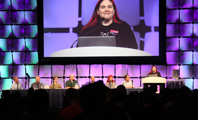 A panel of speakers sits at a table on a stage, with a large screen behind them showing a woman speaking.